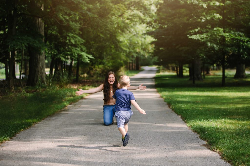 A woman holds open her arms to hug her toddler son on a path through the woods