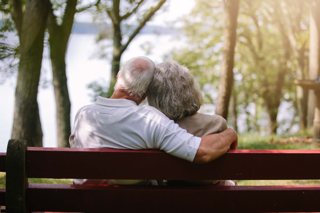 An older couple sits on a bench on the woods together.