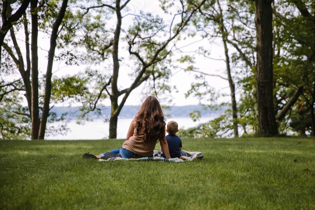 A mom and her toddler son sit on a picnic blanket on the woods.